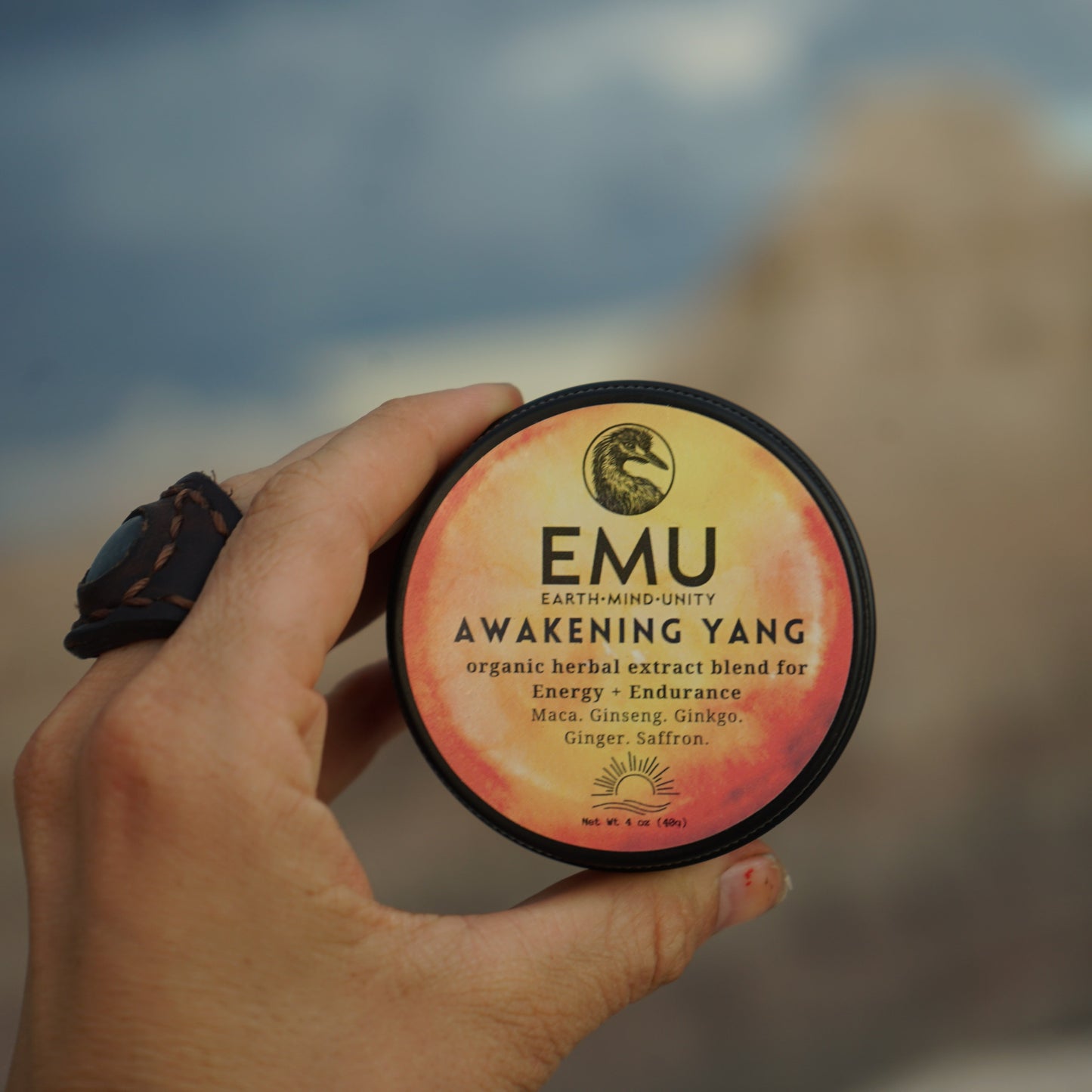 Hand holding a container of EMU Awakening Yang supplement with a blurred natural background