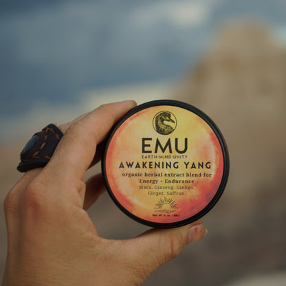 Hand holding a container of EMU Awakening Yang supplement with a blurred natural background